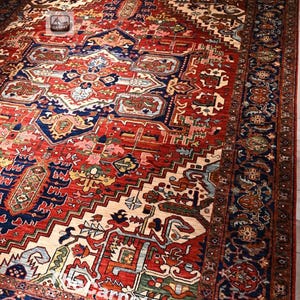 May include: A large rectangular area rug featuring a complex geometric pattern in shades of deep red, navy blue, cream, and green. The design includes a central medallion surrounded by intricate floral and tribal motifs with a wide decorative border.