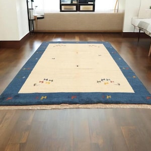 May include: Rectangular rug with a cream-colored center and a deep blue border. The rug features small, colorful geometric patterns and is placed on a dark wood floor. The rug is a decorative floor covering.