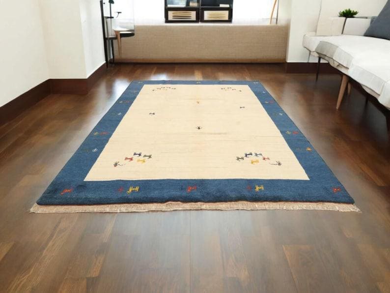 May include: Rectangular rug with a cream-colored center and a deep blue border. The rug features small, colorful geometric patterns and is placed on a dark wood floor. The rug is a decorative floor covering.