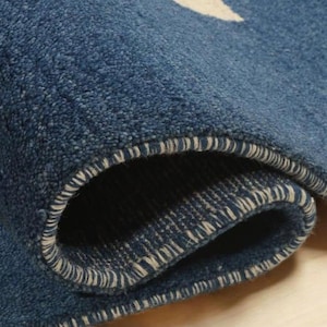 May include: Close-up of a rolled-up blue rug with a white and yellow pattern. The rug has a textured surface and a white stitched border. The rolled section reveals the underside of the rug.
