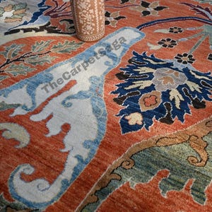 May include: An intricately designed, rust-colored rug with floral and geometric patterns in shades of blue, green, and cream. A decorative, light brown vase with floral cutouts sits atop the rug. The rug's design includes the text "The Carpet Saga".