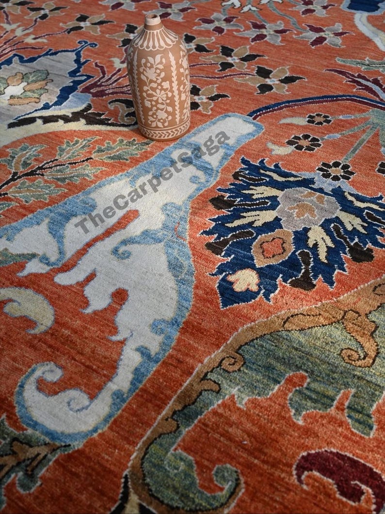 May include: An intricately designed, rust-colored rug with floral and geometric patterns in shades of blue, green, and cream. A decorative, light brown vase with floral cutouts sits atop the rug. The rug's design includes the text "The Carpet Saga".