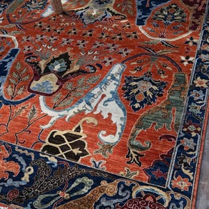 May include: An intricately designed area rug with a burnt orange background and a detailed pattern in shades of blue, cream, and brown. The rug features floral and geometric motifs, with a dark blue border. The rug is a traditional style.