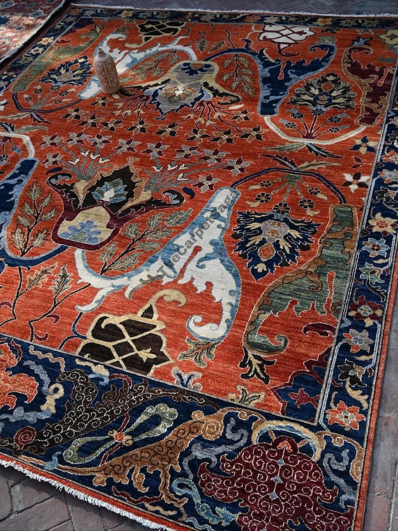 May include: An intricately designed area rug with a burnt orange background and a detailed pattern in shades of blue, cream, and brown. The rug features floral and geometric motifs, with a dark blue border. The rug is a traditional style.