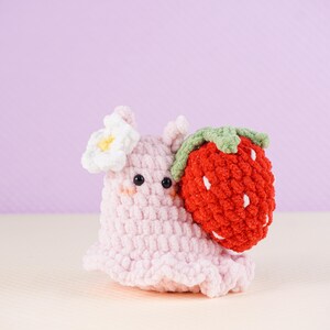 May include: A handmade pink crochet snail with a white flower and a red crochet strawberry. The snail has black eyes and a smiling face. The strawberry has green leaves and white seeds.