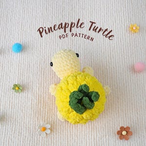 May include: A crocheted pineapple turtle amigurumi toy with a yellow and green pineapple shell. The toy is on a white fuzzy surface with small decorative flowers.