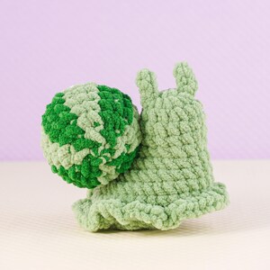 May include: A handmade crochet snail toy in shades of green. The snail's body is a light green, with a ruffled edge. The shell is a sphere of green and light green yarn. The snail has two small antennae.