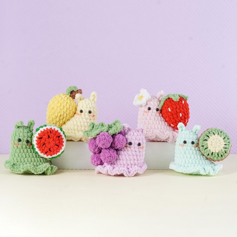 May include: A collection of six crocheted fruit-themed creatures. Each creature is a different color and holds a crocheted fruit, including a watermelon, lemon, grapes, strawberry, and kiwi. The creatures have small black eyes and are set against a light purple background.