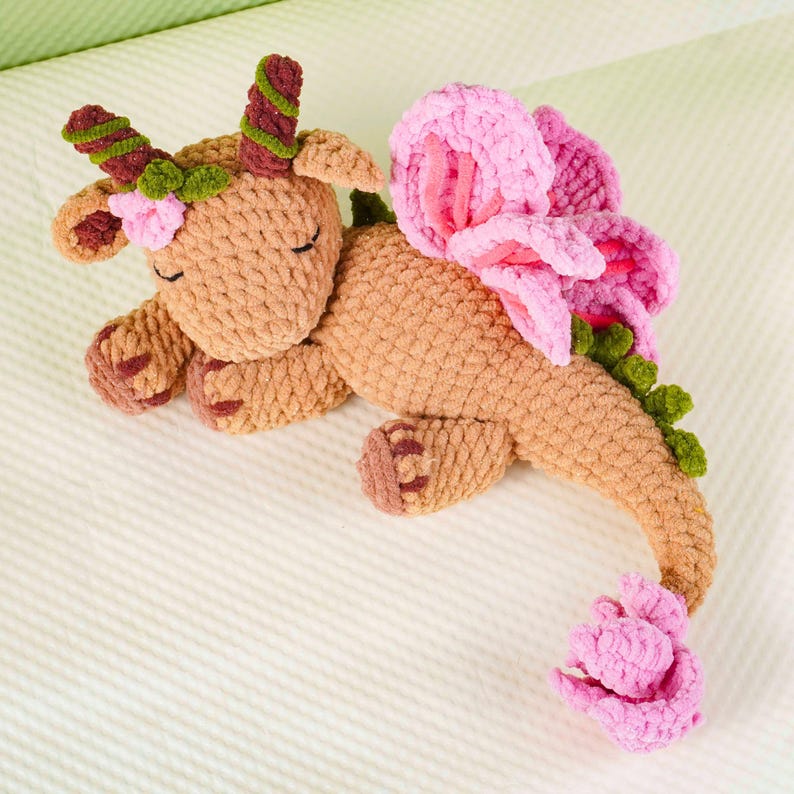 May include: A handmade, plush dragon toy in shades of tan, brown, pink, and green. The dragon features flower-like wings, a flower tail, and a floral crown. The toy is lying down on a white surface.