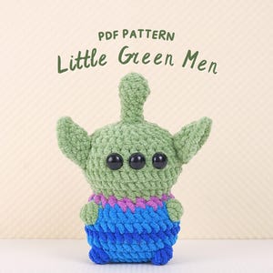 May include: A crocheted green alien with three eyes and a blue and purple striped body. The alien is standing on a white surface. The text "PDF PATTERN Little Green Men" is above the alien.