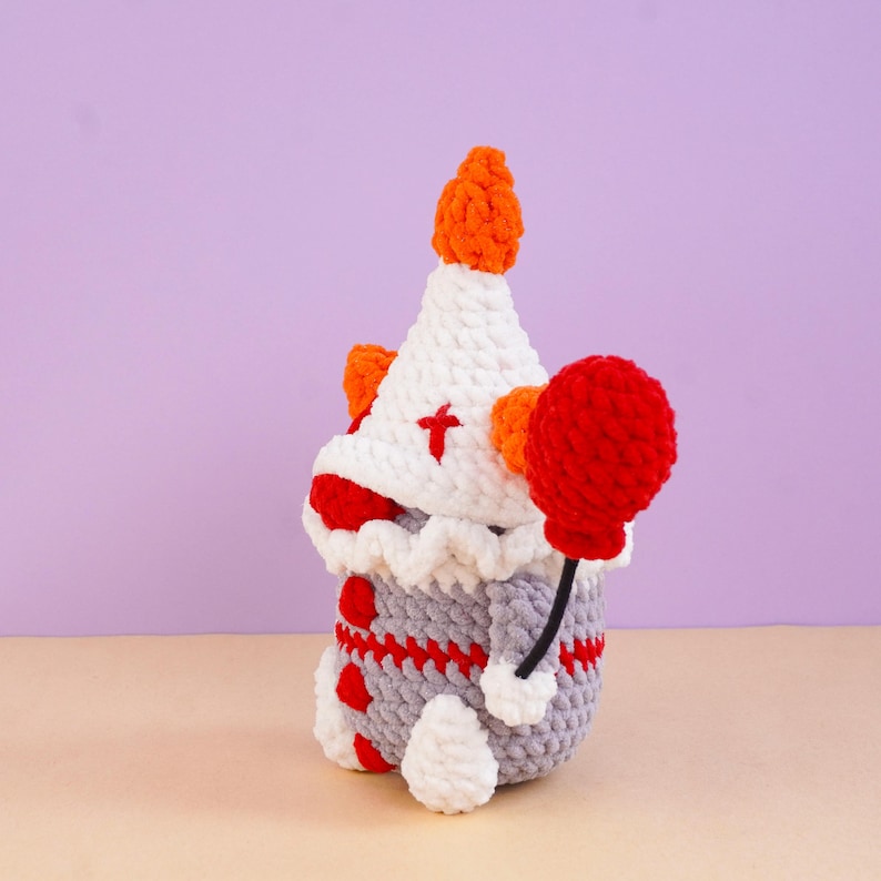 May include: A crocheted clown doll featuring a white pointed hat with an orange pom-pom, a ruffled collar, and a grey body with red accents. The doll holds a bright red balloon on a black string, with red details on the hat and body.