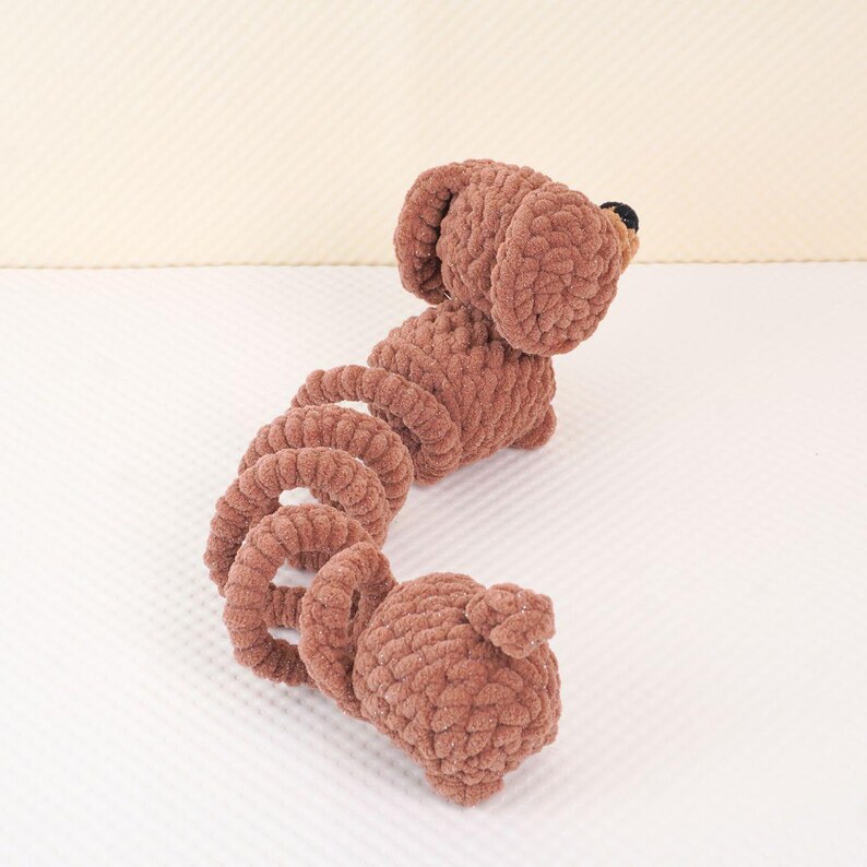 Puede incluir: Un juguete de dachshund de crochet marr&oacute;n con la cola enroscada. El juguete est&aacute; hecho de hilo suave y tiene una expresi&oacute;n linda y juguetona.