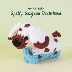 May include: A handmade crochet dachshund dog with white fur, brown spots, and a blue belly. The dog has a black nose and eyes. The text "PDF PATTERN Spotty Sausage Dachshund" is at the top of the image.