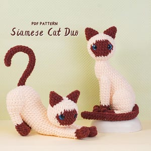 May include: Two crocheted Siamese cats with cream-colored bodies and dark brown ears, faces, paws, and tails. One cat is in a stretching pose with its tail arched, while the other sits upright. The text PDF PATTERN Siamese Cat Duo is visible.