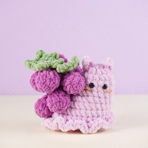 May include: A handmade crochet snail toy in shades of purple and lavender, with a cluster of purple grapes and green leaves on its back. The snail has small black eyes and a cute, smiling expression. The toy is made of soft, textured yarn.