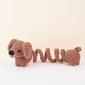Puede incluir: Un juguete de dachshund de crochet marr&oacute;n con una cola larga y rizada. El juguete tiene ojos negros y una nariz negra.