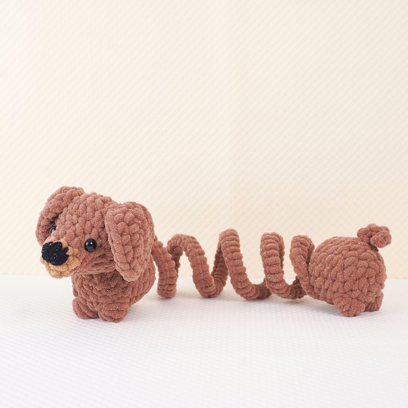 Puede incluir: Un juguete de dachshund de crochet marr&oacute;n con una cola larga y rizada. El juguete tiene ojos negros y una nariz negra.