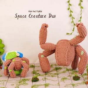 May include: Two crocheted creatures in brown yarn with light green accents. The larger creature has four raised limbs, while the smaller one wears a blue and white striped hat. Text above reads PDF PATTERN Space Creature Duo.