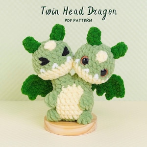 May include: A crochet plush toy featuring a two-headed dragon with light green and cream yarn, dark green wings, and black eyes. The text Twin Head Dragon PDF PATTERN is visible above the creature, which stands on a circular wooden base.