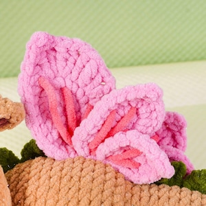 May include: Close-up of a handmade, plush flower with pink petals and red accents. The flower is part of a larger craft project, with a tan base and green leaves visible. The background is a soft green.