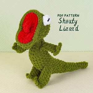 May include: A handmade green crochet lizard with a red mouth and white eye. The lizard is in a standing position with its mouth open. The text "PDF PATTERN Shouty Lizard" is in the upper right corner.
