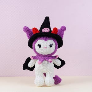 May include: A handmade crochet doll in white, purple, black, and red. The doll is wearing a witch hat with a skull, a purple collar, and black wings. The doll has a cute face and a small tail.