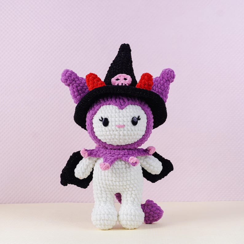 May include: A handmade crochet doll in white, purple, black, and red. The doll is wearing a witch hat with a skull, a purple collar, and black wings. The doll has a cute face and a small tail.