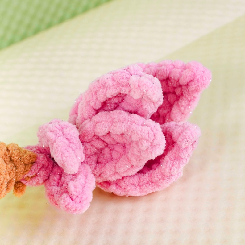 May include: Close-up of a handmade pink crochet flower with textured petals. The flower is made of soft, fluffy yarn and has a detailed, three-dimensional design. The stem is a light brown color.