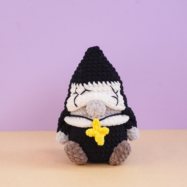 May include: A crocheted plush figure featuring a black pointed hat, a white face with closed eyes, a gray nose, and a black body. The figure wears a white collar and a bright yellow cross-shaped pendant. It has gray hands and feet.