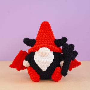May include: A crocheted gnome figure featuring a tall red pointed hat, black devil horns, and black bat wings. The gnome has a white beard, a round nose, and holds a black three-pronged pitchfork. The body is black with red accents.