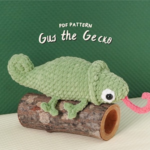 May include: A handmade green crocheted gecko toy with a pink tongue, perched on a piece of wood. The gecko has black and white eyes and is made of a soft, textured yarn. The text "PDF PATTERN Gus the Gecko" is above the gecko.