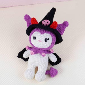 May include: A handmade crochet doll in white, purple, black, and red. The doll has a witch hat with a skull, purple ears, and a purple collar with pink accents. The doll has black wings and a purple tail.