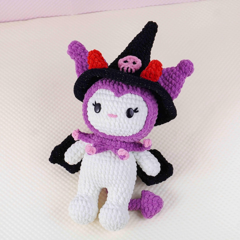 May include: A handmade crochet doll in white, purple, black, and red. The doll has a witch hat with a skull, purple ears, and a purple collar with pink accents. The doll has black wings and a purple tail.
