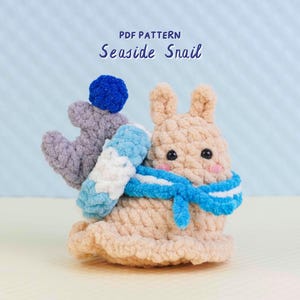 Könnte beinhalten: Ein handgefertigtes gehäkeltes Schneckenspielzeug in Beige, mit einem blau-weißen Schal und einem blauen Bommel. Die Schnecke hat schwarze Augen und rosa Wangen. Der Text "PDF PATTERN Seaside Snail" befindet sich oben im Bild.