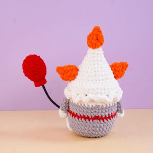 May include: A crocheted figure featuring a white pointed hat with an orange top, orange side tufts, and a gray body with a red horizontal stripe. A thin black wire extends from the side, holding a small, textured red balloon shape.