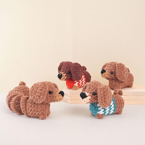Puede incluir: Cuatro juguetes de peluche de dachshund tejidos a crochet en marr&oacute;n, con su&eacute;teres de diferentes colores. Los su&eacute;teres son rojo y blanco, azul y blanco, y azul verdoso y blanco.