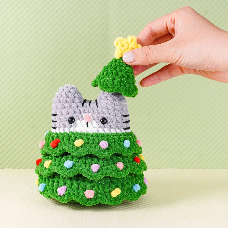 Peut inclure: Un sapin de No&euml;l en crochet fait &agrave; la main en forme de chat. Le corps est vert, la t&ecirc;te grise, avec des ornements color&eacute;s. Une main place un petit arbre vert avec une &eacute;toile jaune. Le chat a des rayures noires et un visage blanc et rose.