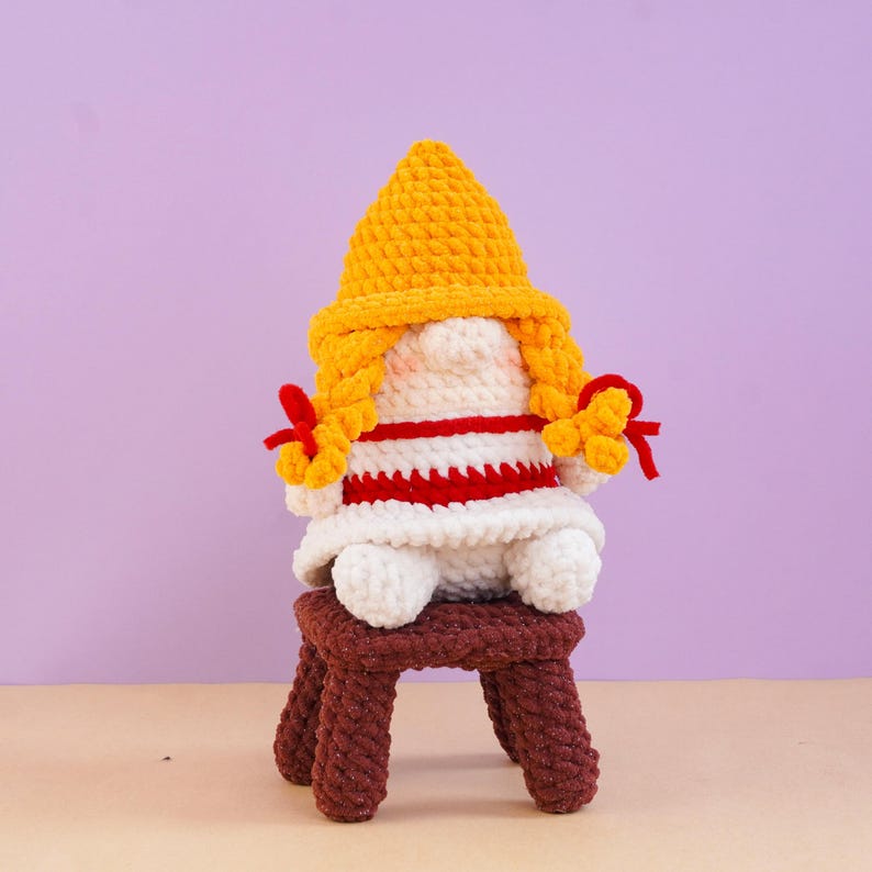May include: A crocheted gnome figure with a bright yellow pointed hat, long yellow braided hair, and a white body featuring horizontal red stripes. The gnome sits on a small, dark brown crocheted stool, with red ribbons tied at the ends of its braids.