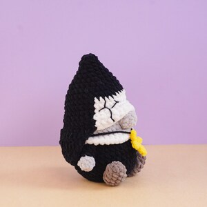 May include: A crocheted plush figure featuring a black pointed hood, a white face with stitched closed eyes, and a grey nose. The figure has a black body, small grey hands, and brown feet, and it holds a small yellow star-shaped accessory.