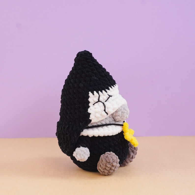 May include: A crocheted plush figure featuring a black pointed hood, a white face with stitched closed eyes, and a grey nose. The figure has a black body, small grey hands, and brown feet, and it holds a small yellow star-shaped accessory.