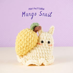 Puede incluir: Un juguete de ganchillo hecho a mano con forma de caracol, con una concha amarilla en forma de mango y un cuerpo color crema. El caracol tiene pequeños ojos negros y mejillas rosadas. El texto "PDF PATTERN Mango Snail" está encima del juguete.