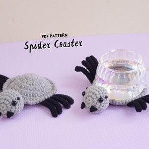 May include: Two crochet spider coasters in gray and black yarn. Each coaster features a round gray body, a small gray head with black bead eyes, and eight black legs. One coaster supports a clear glass mug. Text reads: PDF PATTERN Spider Coaster.