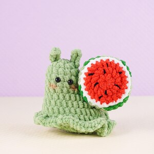 May include: A handmade green crochet snail with a watermelon shell. The snail has black eyes and blush on its cheeks. The watermelon shell is red with black seeds, white rind, and green edges. The snail is on a light yellow surface against a purple background.