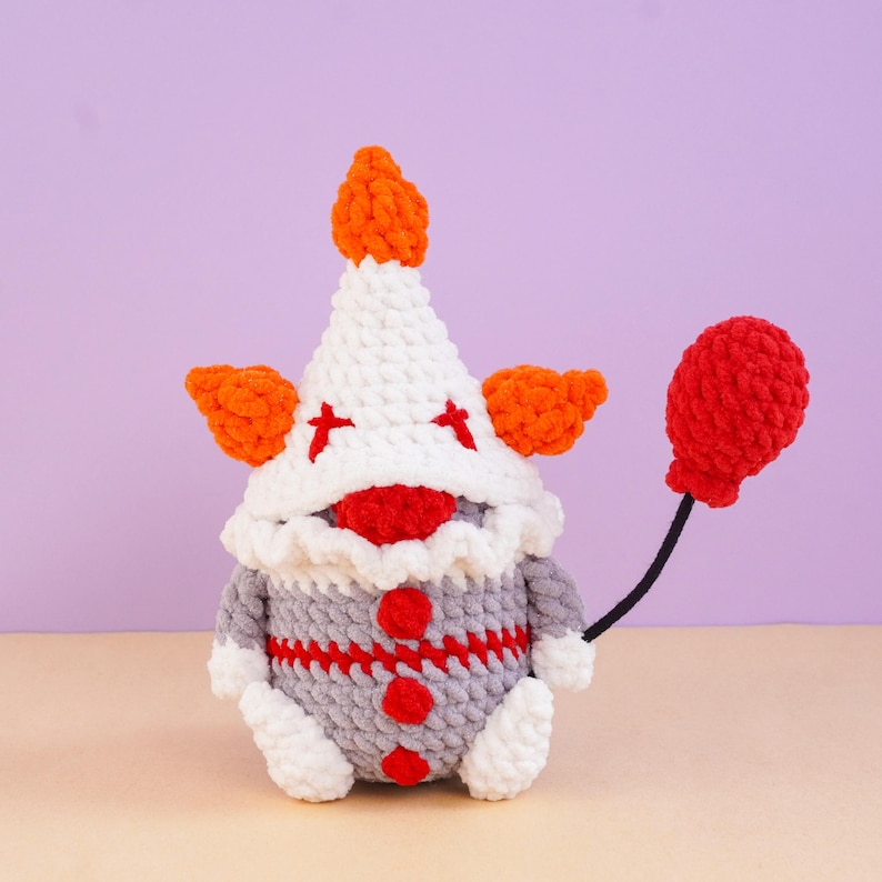 May include: A crocheted clown gnome figure featuring a white pointed hat with orange tufts, a red nose, and a grey body with three red buttons. The figure holds a red crocheted balloon on a thin black string, with red accents on the hat.