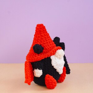 May include: A crocheted gnome figure featuring a tall, textured red hat with black accents, a round black body, and a white crocheted beard. The gnome has small white hands and red feet, crafted from soft yarn with a visible, chunky stitch pattern throughout.