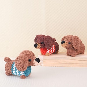 Puede incluir: Tres juguetes de perro salchicha de crochet marr&oacute;n con su&eacute;teres de diferentes colores. El dachshund de la izquierda lleva un su&eacute;ter azul y blanco, el dachshund del medio lleva un su&eacute;ter rojo y blanco y el dachshund de la derecha lleva un su&eacute;ter marr&oacute;n.