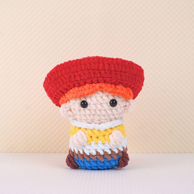 May include: A crocheted toy figure of a person wearing a red hat, orange hair, a yellow shirt, white pants, and blue shoes.