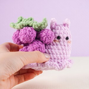 May include: A handmade crochet snail toy in shades of purple, with a cluster of purple grapes and green leaves on its back. The snail has black eyes and a smiling expression. The toy is held in a hand against a light purple background.