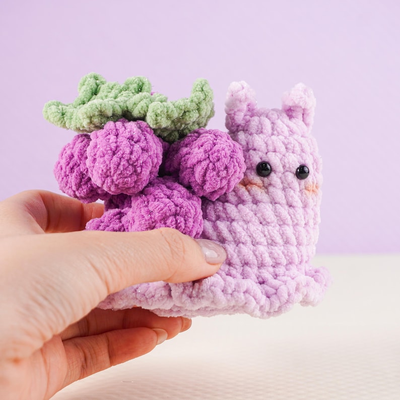 May include: A handmade crochet snail toy in shades of purple, with a cluster of purple grapes and green leaves on its back. The snail has black eyes and a smiling expression. The toy is held in a hand against a light purple background.