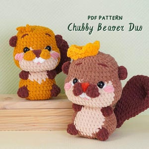 May include: Two crocheted beaver plushies in shades of brown, tan, and orange sit on a wooden surface. The plushies have small black eyes, pink cheeks, and a yellow crown. The text "PDF PATTERN" and "Chubby Beaver Duo" are visible.